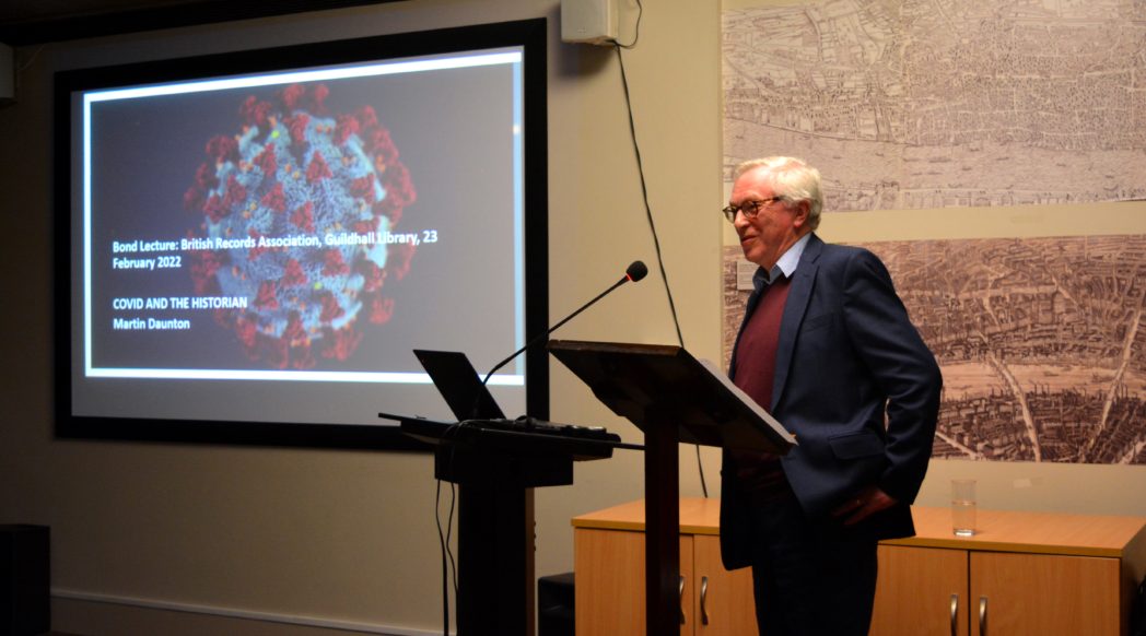 a man stood at a lecturn with a powerpoint display behind him
