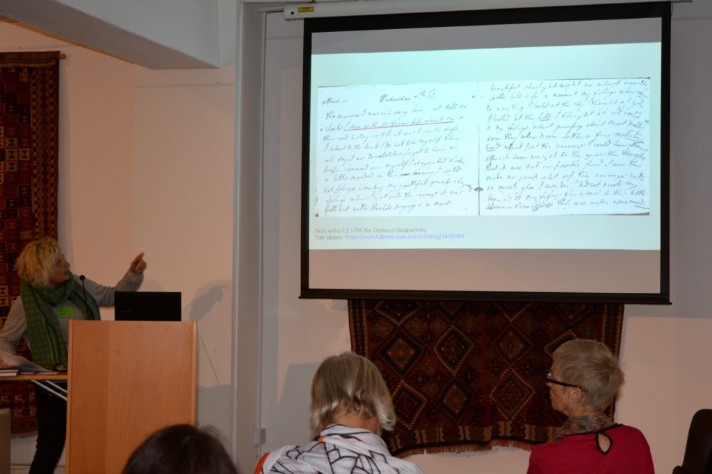 A woman stood at a lectern with a laptop on it, giving a presentation. A projection screen is to her left and she is pointing to a manuscript shown on it