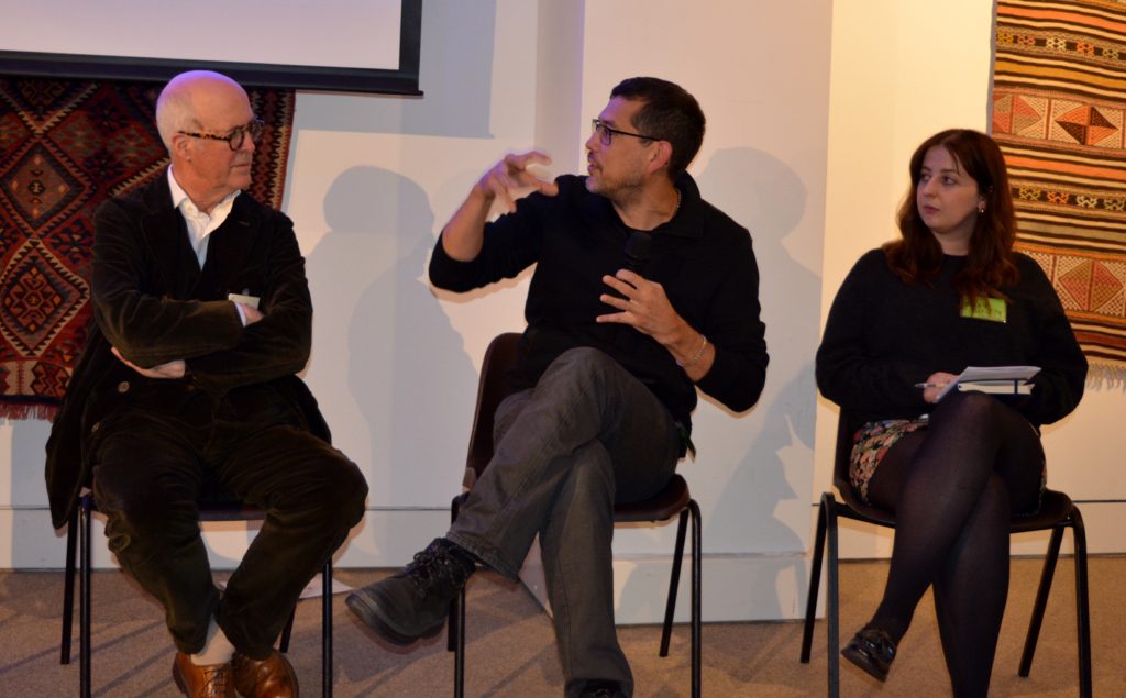 A panel of three individuals sat in chairs in a row. The centre man is gesticulating as he talks