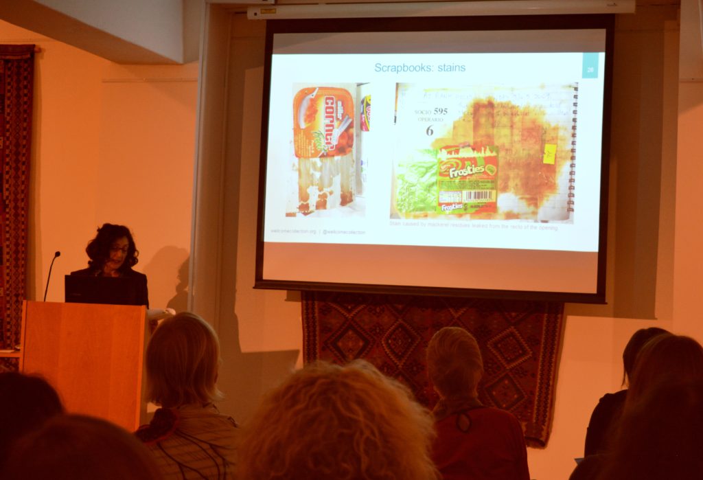 A woman stood at a lectern with a laptop on it, giving a presentation. A projection screen is to her left showing a yogurt carton conserved by her, pasted in a scrapbook